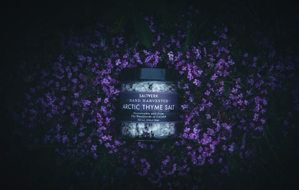Jar of Arctic Thyme Salt surrounded by purple flowers on a dark background