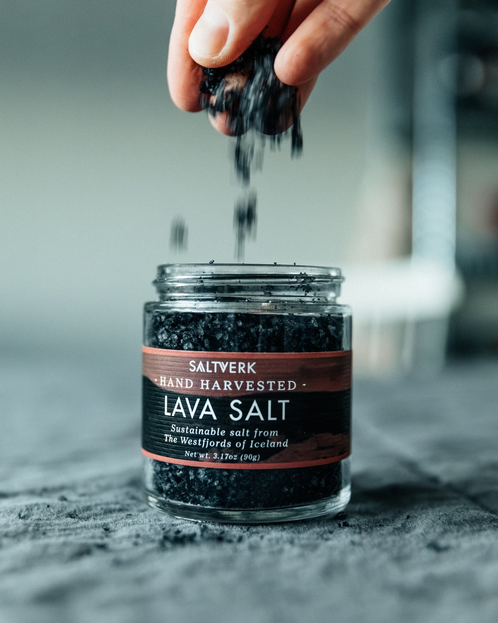 Jar of Saltverk lava salt with a hand sprinkling salt over it.